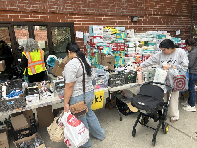 Utility tables are organized with bins, boxes and crates of toiletries, such as baby wipes, diapers and toothpaste. Several people are holding plastic bags and browsing through the available items. 