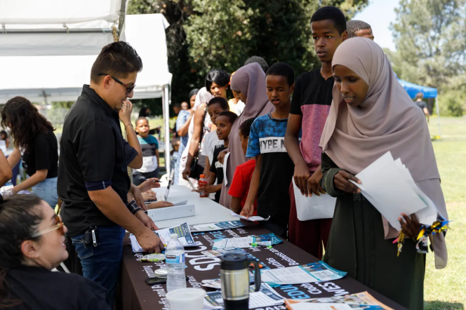 A line of young people, most with dark brown skin, stand in front a display table stffed by a man wearing sunglasses and a black shirt.