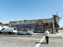 A man wearing a beige jacket, dark pants and a red baseball cap crossing a street. He is walking towards a building with a mural of people painted on the side with the words "herbs of Mexico. Herbs from around the world."