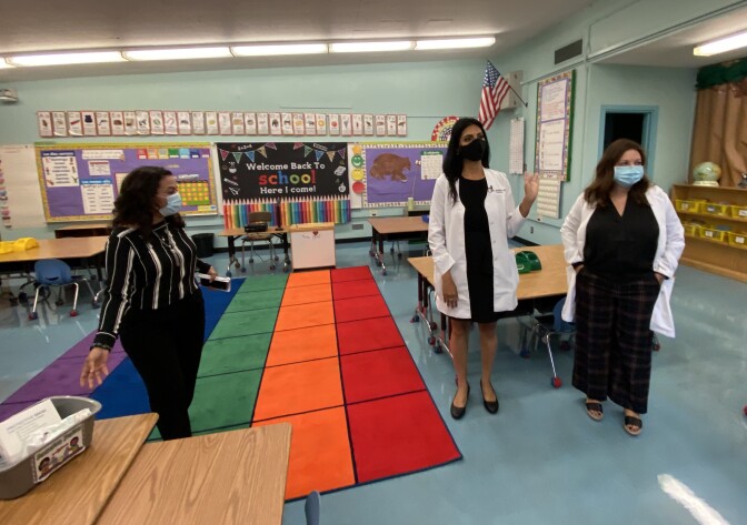 Three school officials, including two doctors in white lab coats, stand in a classroom with desks and chairs behind them.