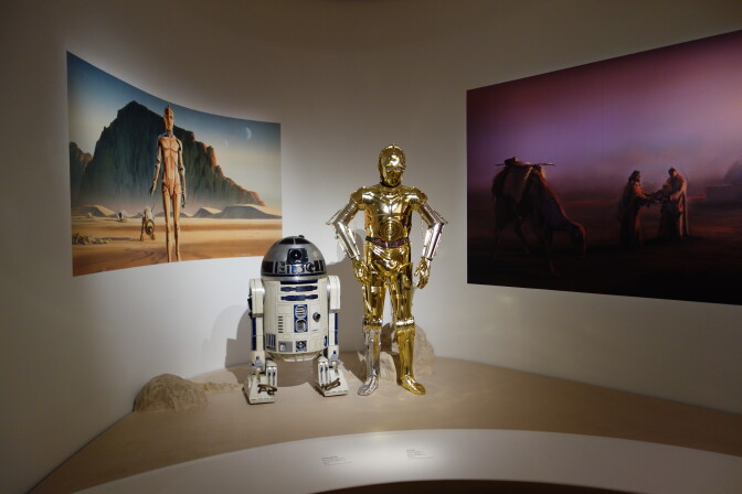Two droids - props from the Star Wars movies - are set up in a museum exhibit with concept art depicting them in the background.