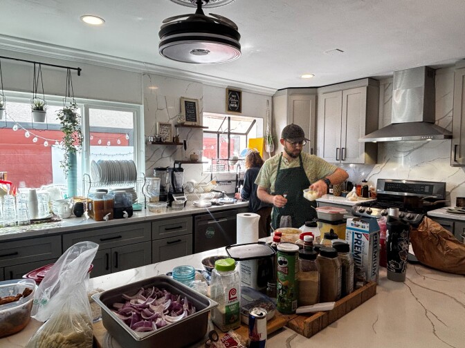 A man in a green apron works at a home kitchen counter surrounded by ingredients including sliced red onions, spices, and condiments. A woman works at the sink in the background. String lights and plants are visible through a window overlooking the backyard dining area.