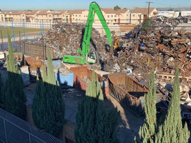 A heavy construction vehicle picks up trash and metal as it stands in two large piles almost as tall as it with a high school in the background.