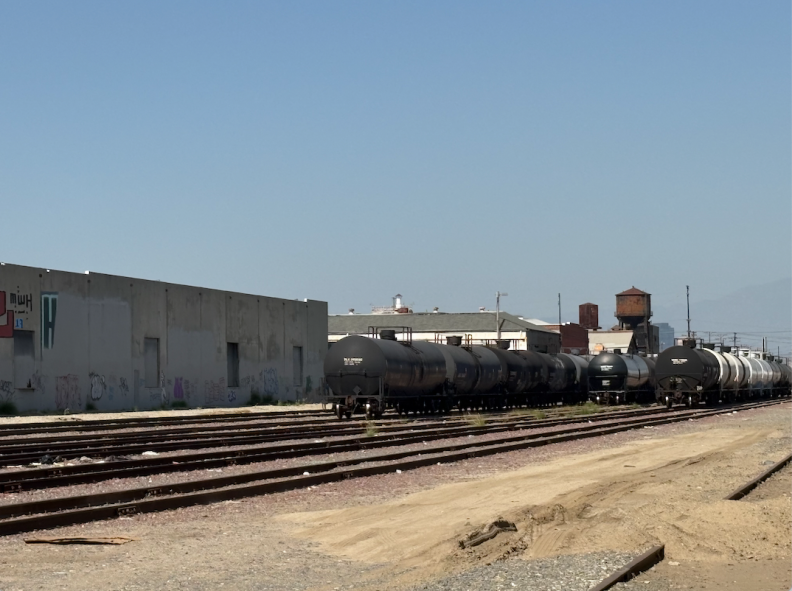 Freight trains idle on a track. There is light brown dirt around the trains, and white buildings in the background.