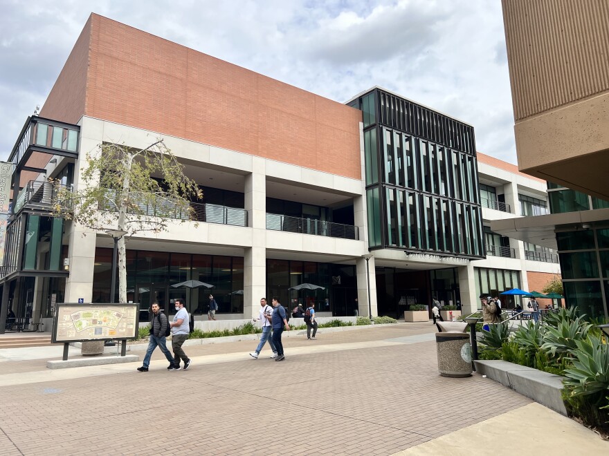 Six students are seen walking, some in pairs, to their next class at East Los Angeles College. They are walking past a multi-story building made of brick with large glass windows.