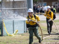 A women wearing a yellow shirt, green pants and a white hard hat holds and sprays a yellow fire hose. 