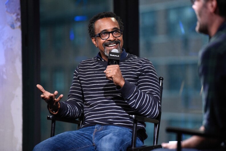 Comedian Tim Meadows sits in a chair holding a mic and wearing a black and white striped shirt and jeans.