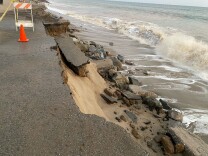 The side of the roadway has shattered and dropped into the sand as waves roll to shore. A caution cone and hazard sign are in the roadway.