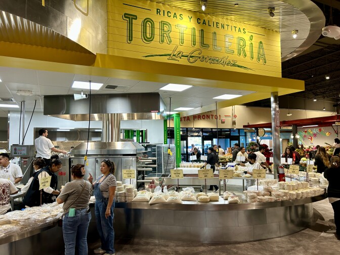 A wide shot of a long curving counter stacked with tortillas and masa for making tortillas. There are customers standing around the outside, and workers manning large metal machines on the inside. Above the area a bright yellow sign reads "Tortillería La González."