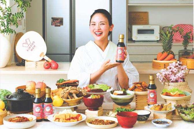 A light-skinned woman wearing a white kimono smiles while holding a bottle of soy sauce. in front of her on a counter is a large spread of food. 