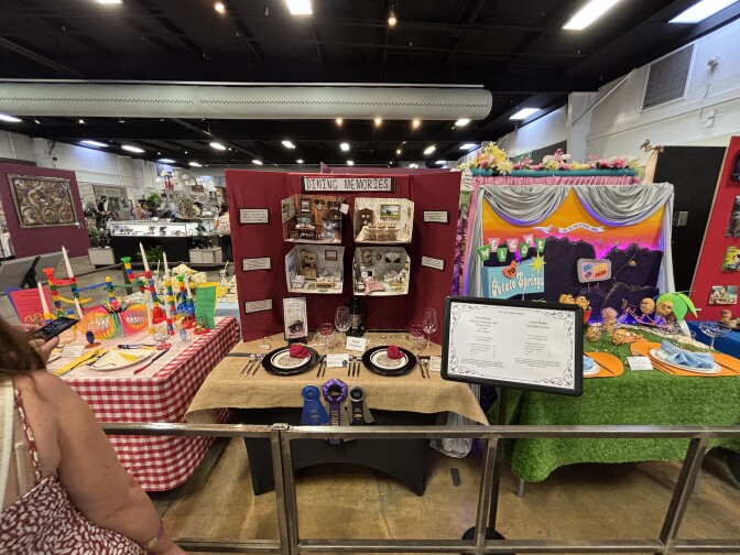 A row of three highly-decorated mini table setting displays. The one on the left has a red gingham table cloth and childrens' toys as centerpieces, the center one features a back display with dollhouse-sized minatures, and the one on the right reads "Welcome to Potato Springs" with potatoes acting out different scenes on an astroturf tablecloth.