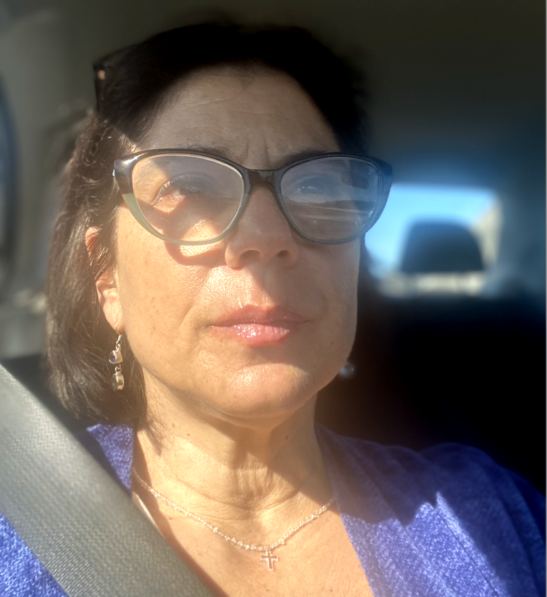 A woman with dark hair sits in a car looking past the camera.