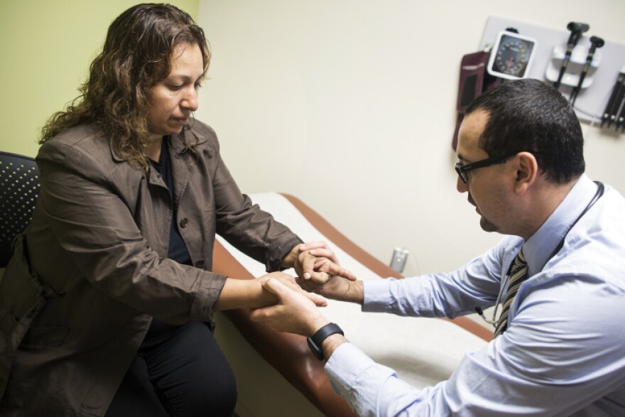 A doctor in a collared shirt and tie, but no coat, holds s a woman's hands. An examining table is behind them.
