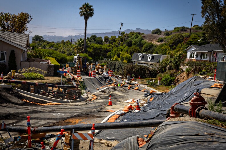 A road passing through a residential neighborhood is damaged due to land movement underneath. Orange safety cones and barriers dot the foreground and black tarps are spread over the area where the road was.