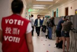 An out of focus person with a red vest that says "vote" in multiple languages looks on as several people stand in voting kiosks in a long hallway.