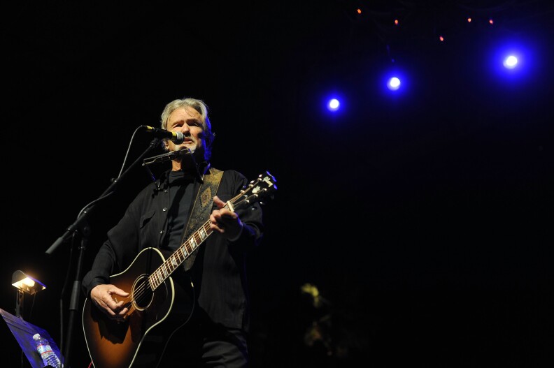 Image shows Kris Kristofferson plays guitar and stands in front of a microphone on stage