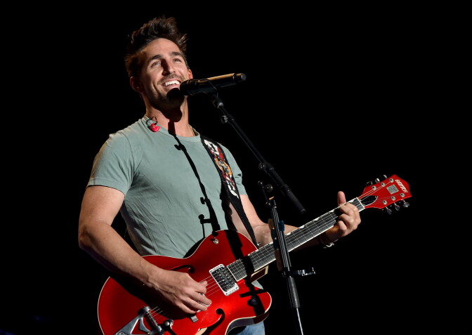 Country singer Jake Owen, a 20- or 30-something white man, smiles while standing onstage in front of a microphone. He holds a bright red guitar.