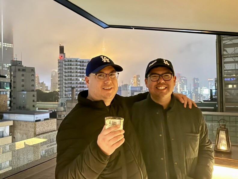 Two men in their 30s wearing blue Dodger hats pose in front of a window overlooking a cityscape.