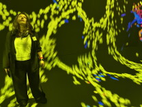 A woman standing in front of an art exhibition comprised of hundreds of yellow-green dots, interspersed with occasional blue dots.