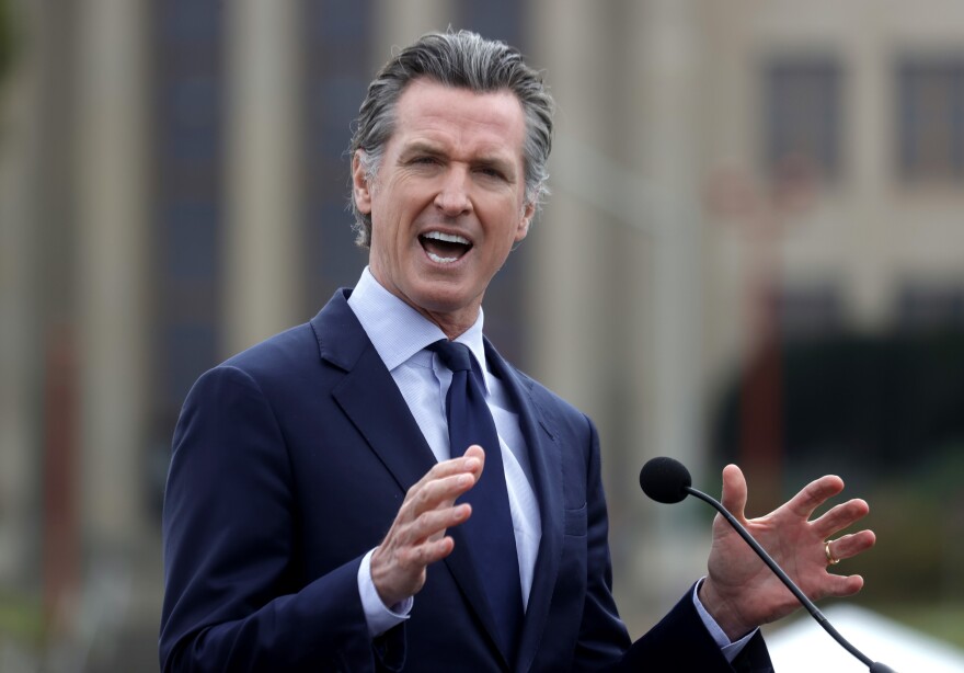 California Governor Gavin Newsom speaks at a podium, his hands raised next to the microphone.