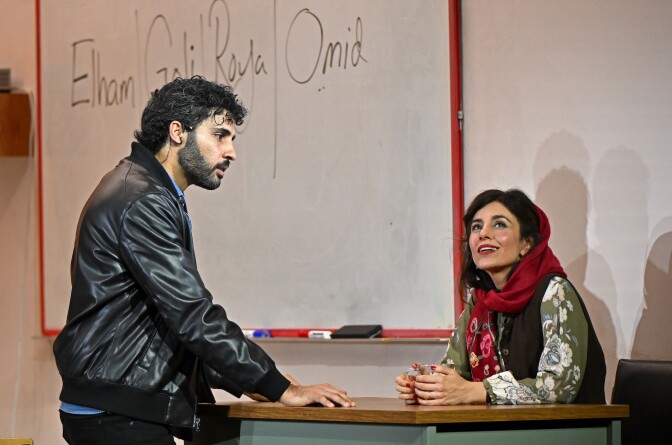 A medium-light-skin-toned man in a black leather jacket stands in front of a desk and looks at a medium-light-skin-toned woman wearing a red head scarf and seated behind the desk. 