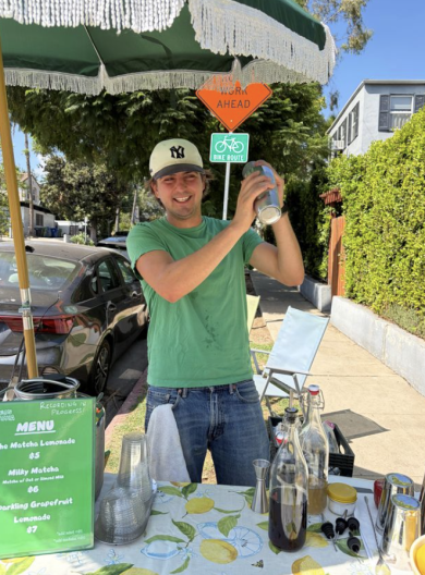 A light skinned man shakes a cocktail shaker at an outdoor drink stand with a menu listing matcha lemonade and sparkling grapefruit lemonade.