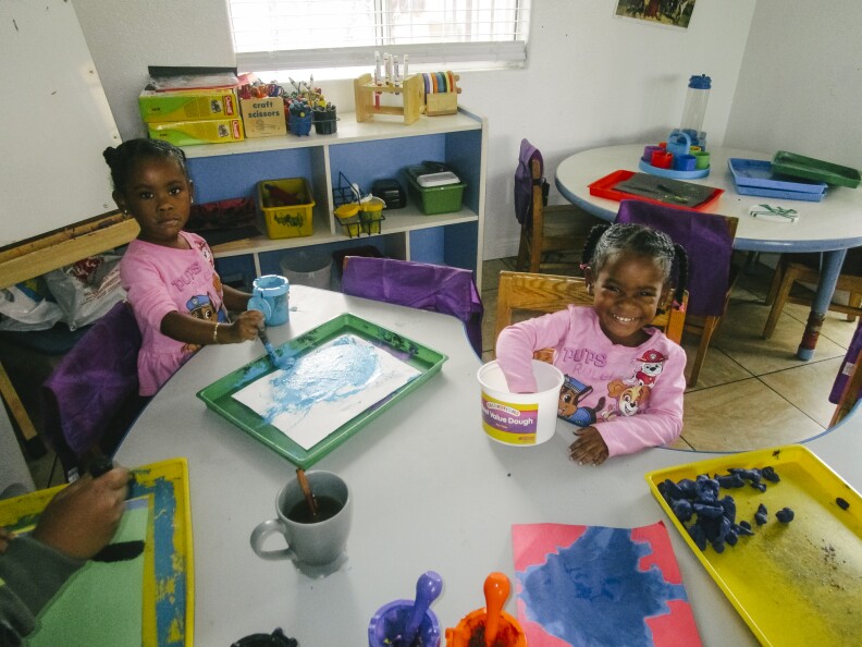 In this image, two children play with paints and play dough at a table in the classroom.