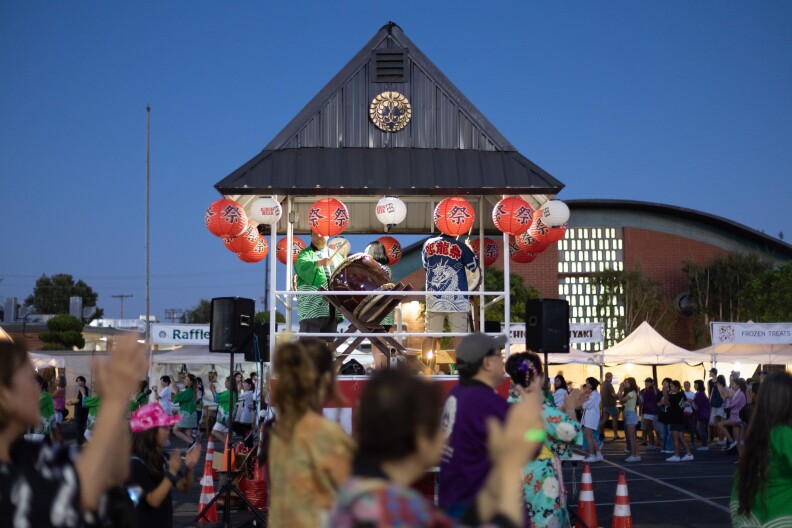 A structure with a pitched tower and Buddhist crest stands in the middle of a dancing crowd. It glows in the evening light.