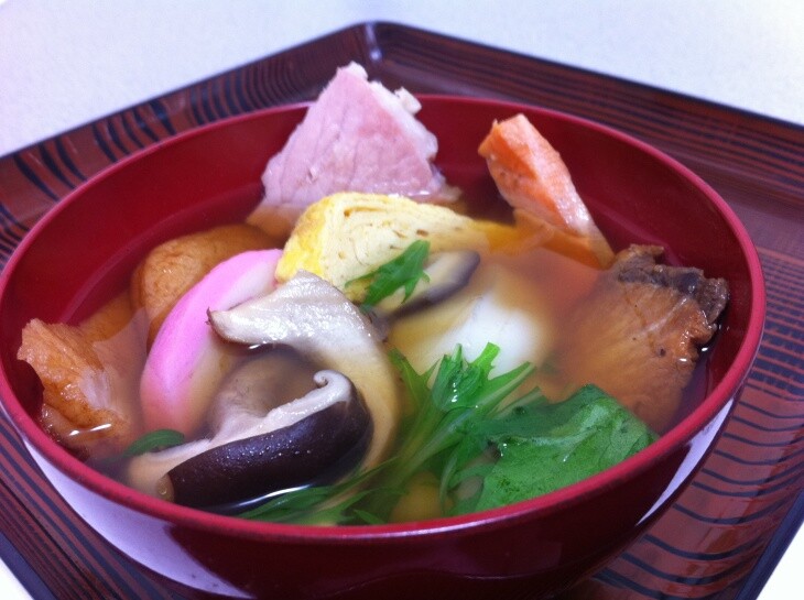A close-up photograph of a red plastic bowl containing a broth so clear that you can easily observe the ingredients inside, including mushrooms, veggies and rice cake.