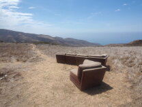 A large light brown sectional, in two parks, has it's seats pointed toward a panoramic view of the ocean and mountains.