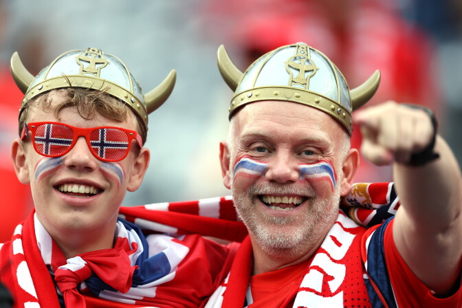 Two male presenting persons are outfitted in Norwegian colors, with red white and blue stripes on their faces, and are wear Viking hats with horns.