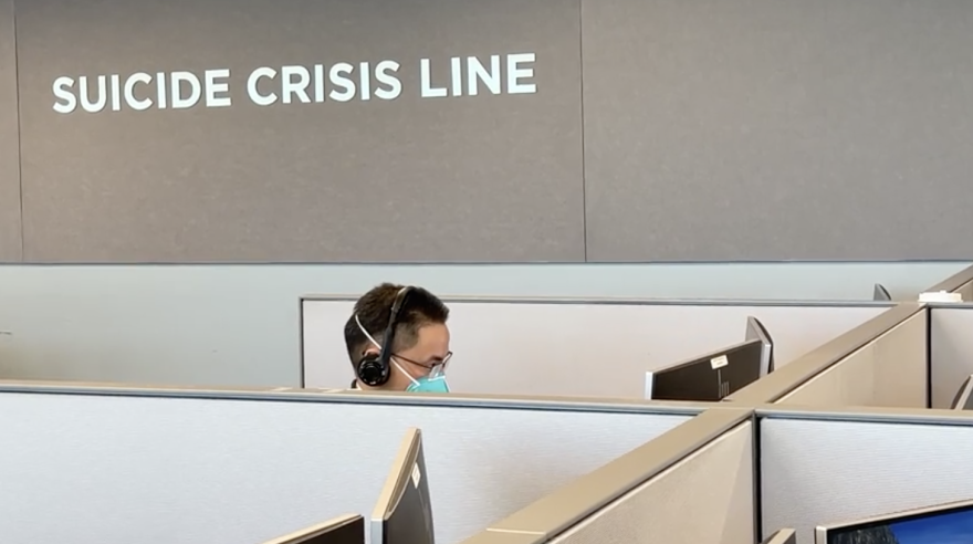 A suicide crisis call center worker sits at a desk wearing a mask. Above him a sign reads "Suicide Crisis Line"