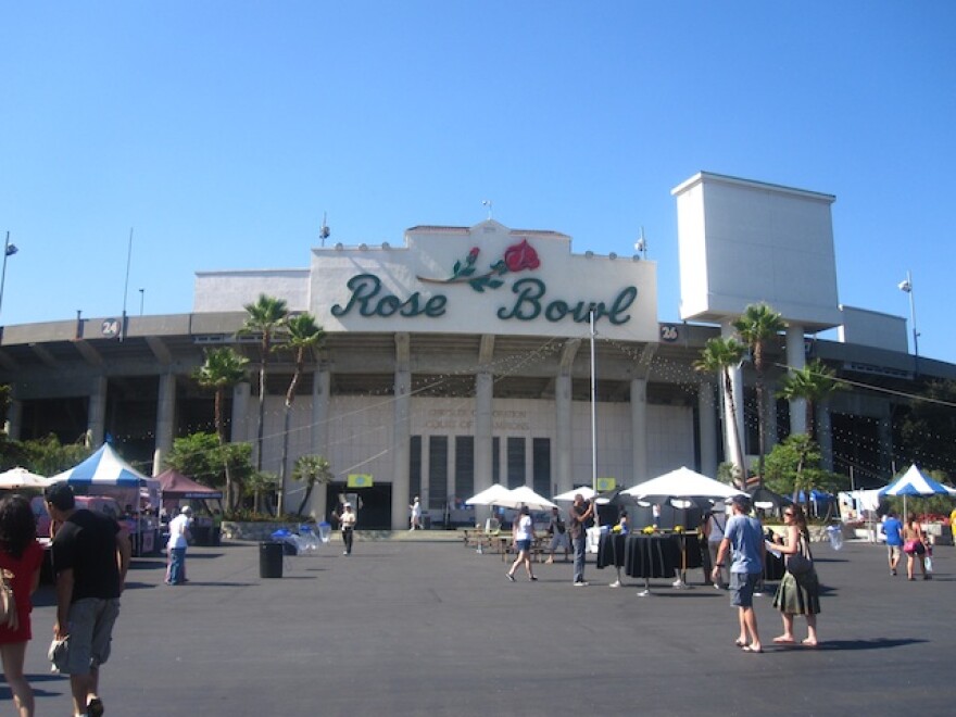 An exterior of the Rose Bowl stadium in Pasadena. White umbrellas dot the parking lot as some people walk towards the main entrance, which depicts a large rose above the words Rose Bowl in cursive lettering.