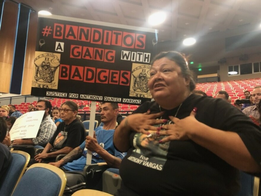 A woman with medium-tone skin clutches her chest as she speak in a room. Behind her a man holds a sign that reads: Banditos a gang with badges