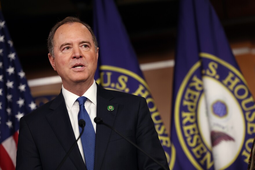 A man with light-tone skin wears a dark suit and blue tie. He stands in front of a mic with flags for the U.S. and U.S. House of Representatives behind him.