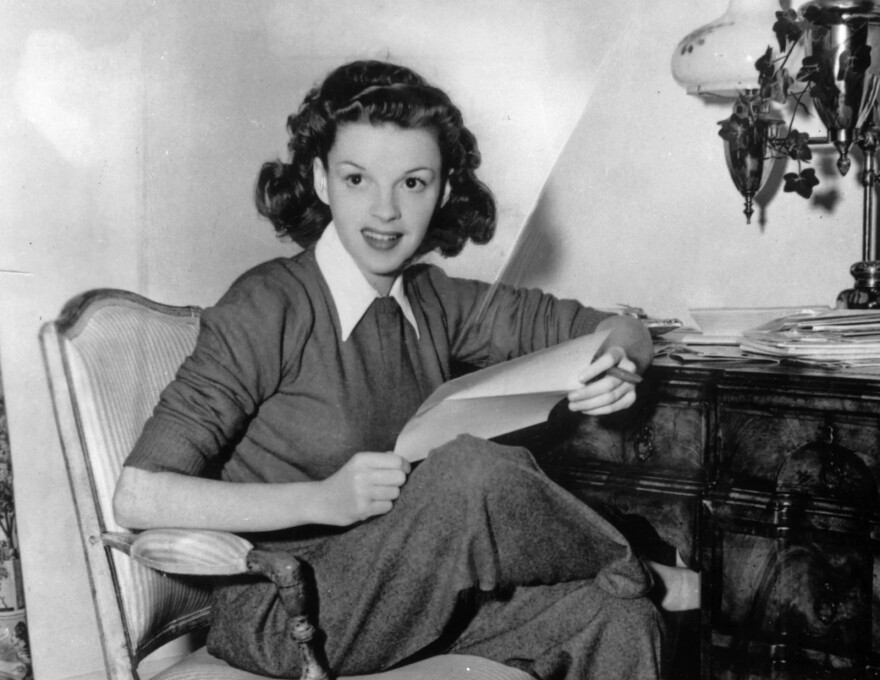 A black and white photo of Judy Garland. She's smiling, sitting in a chair and holding some papers.