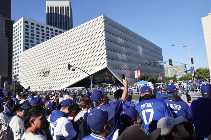 A crowd of people wearing blue Dodgers gear wait on the side of a road with tall buildings in the background.