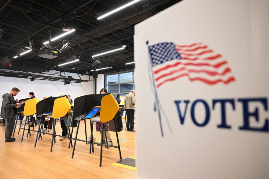 People cast ballots on electronic voting machines. In the foreground is a white voting both with the word "vote" and a U.S. flag printed on the side.