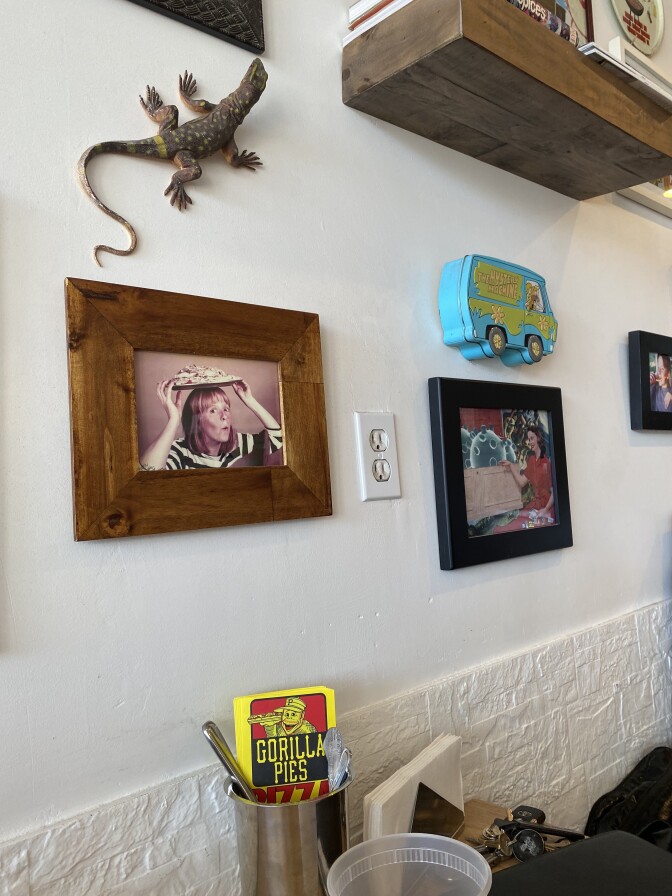 A white wall holds different framed photographs. There is also a toy lizard on the wall. Towards the bottom is a small metal container holding a yellow menu for Gorilla Pies