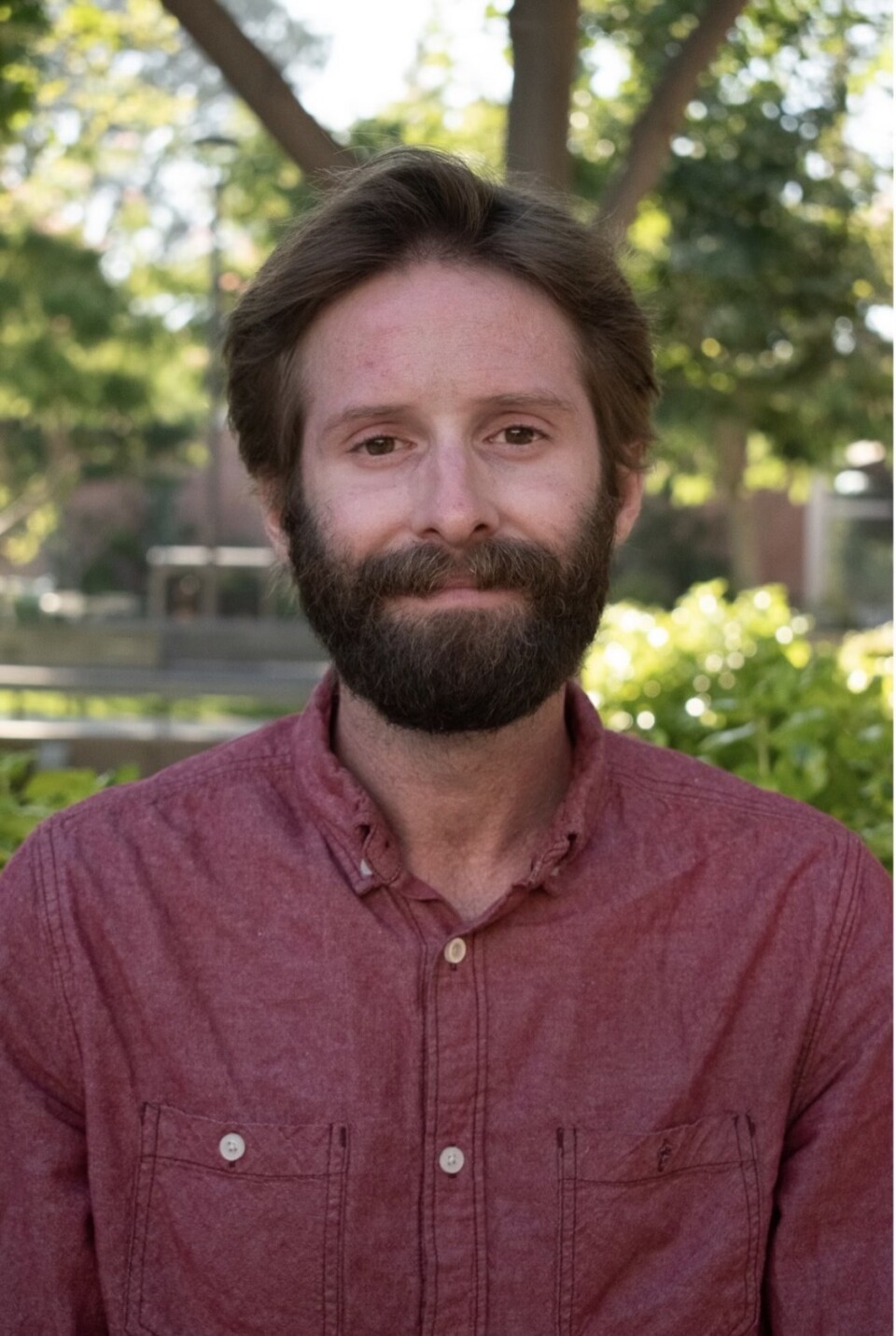 A fair-skinned man with brown hair and a beard wearing a red button-up shirt. Trees and bushes are in the background.