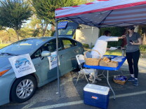 Two women stand under a pop-up shade printed with the American flag. Beside them is a car with several posters hanging from it that say "Canyon Democrats."