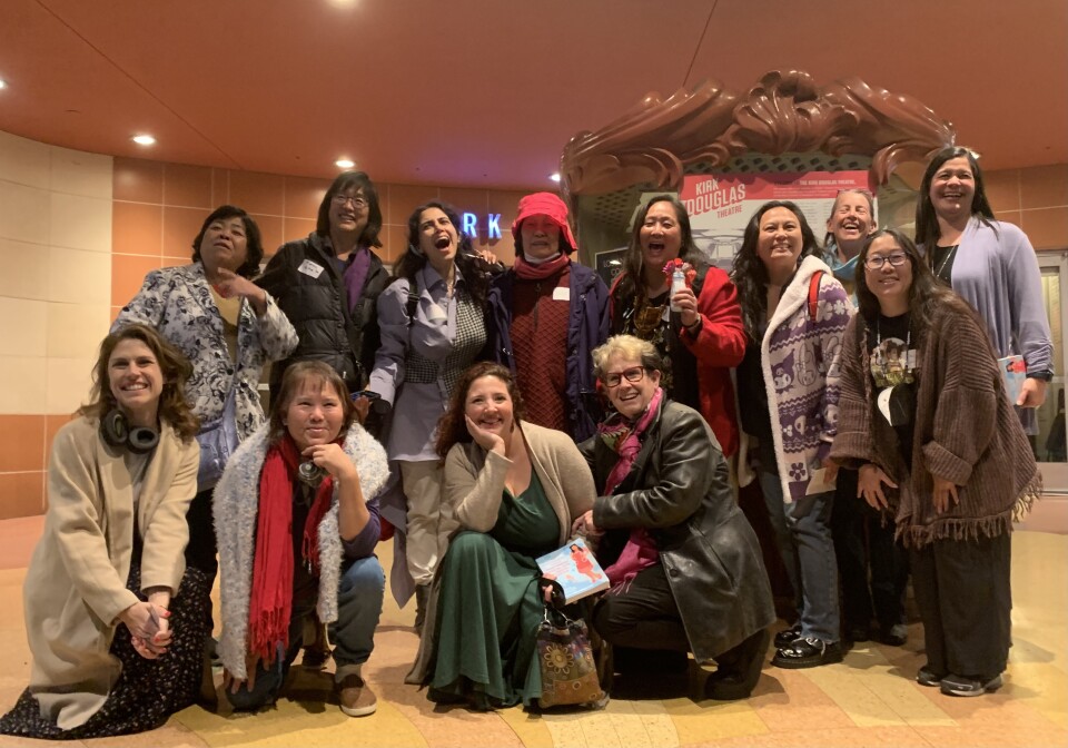 Eleven women of different races pose in front of a theater box office, some standing, some kneeling. 