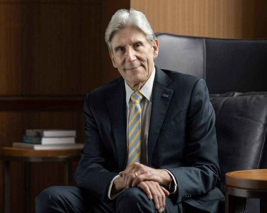 An older man with light skin tone and grey hair sits for a portrait in a black leather chair, wearing a suit and tie.
