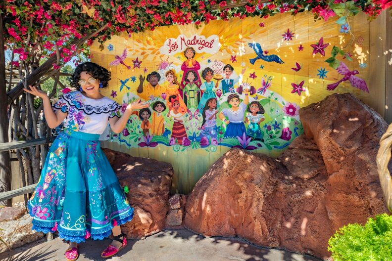 An actress portraying Mirabel from Encanto smiles in from of a Los Madrigal mural, depicting the members of her fictional family.