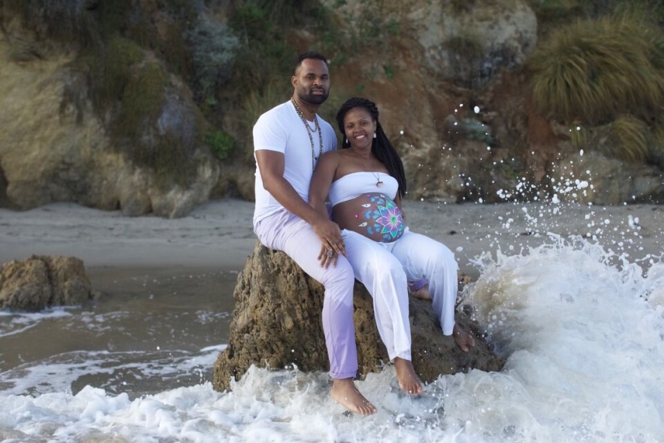 Kacie Blackman sits in front of her husband on a rock with waves splashing at their feet, both wear white and her pregnant stomach has decorative paint on it.