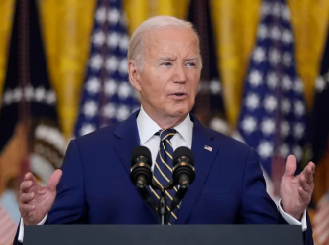 A man with white hair is wearing a blue suit jacket, white shirt, and a striped tie with an American flag pin. He has both hands open and is speaking behind a podium behind a mic with flags in the background. 