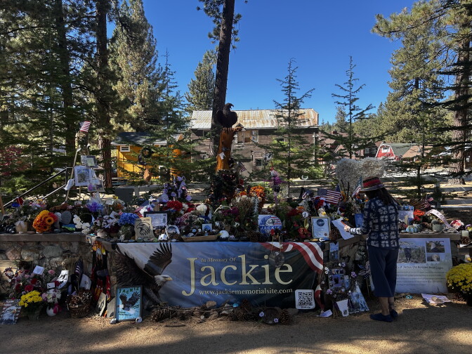 A memorial for Jackie the bald eagle holds flowers, flags, and photos around a banner reading 'In Memory Of Jackie' as a person takes a phone photo of a note.