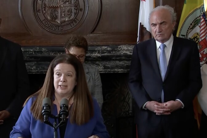 City Attorney Hydee Feldstein Soto, a woman with light skin tone, straight brown hair, wearing a blue blazer stands at a podium and speaks into a microphone. Behind her is Mark Adams, a man with light skin tone, white hair, wearing a navy blue blazer, white shirt, and light blue tie. He stands in front of two flags. 