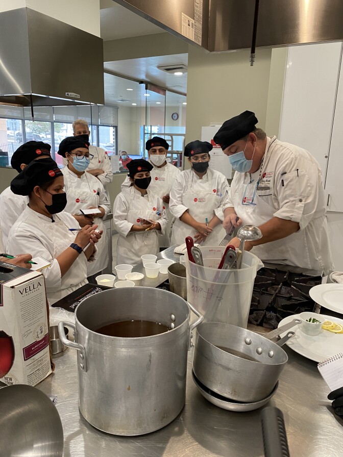 A group of students in chef's aprons, holding notepads and pens, huddles around instructor Danny Harold, watching him cook something on the stove at the Hospitality Training Academy
