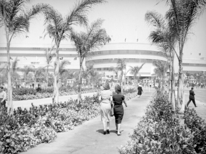 Two women walk together towards Hollywood Park along a path lined with palms. The flags above the building might indicate that it is near the time that the park opened in June, 1938. The clubhouse is the circular building on the right. 